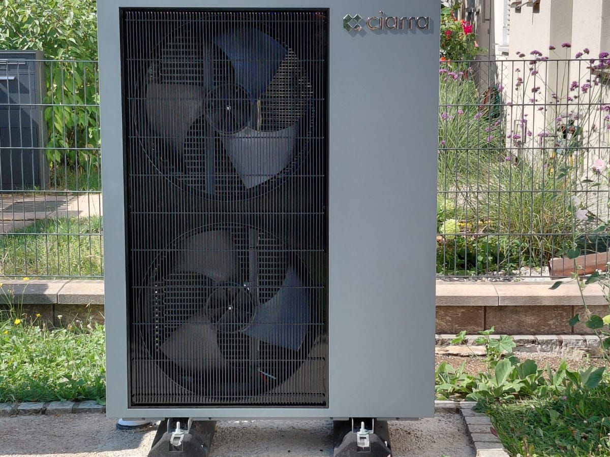 Frontansicht der Außeneinheit mit zwei übereinanderliegenden Ventilatoren, dahinter Gartenzaun und Beet